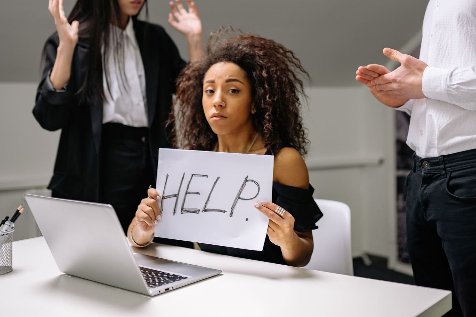 Femme débordée tenant une feuille sur laquelle est écrit « Help », entourée de collègues qui la sollicitent